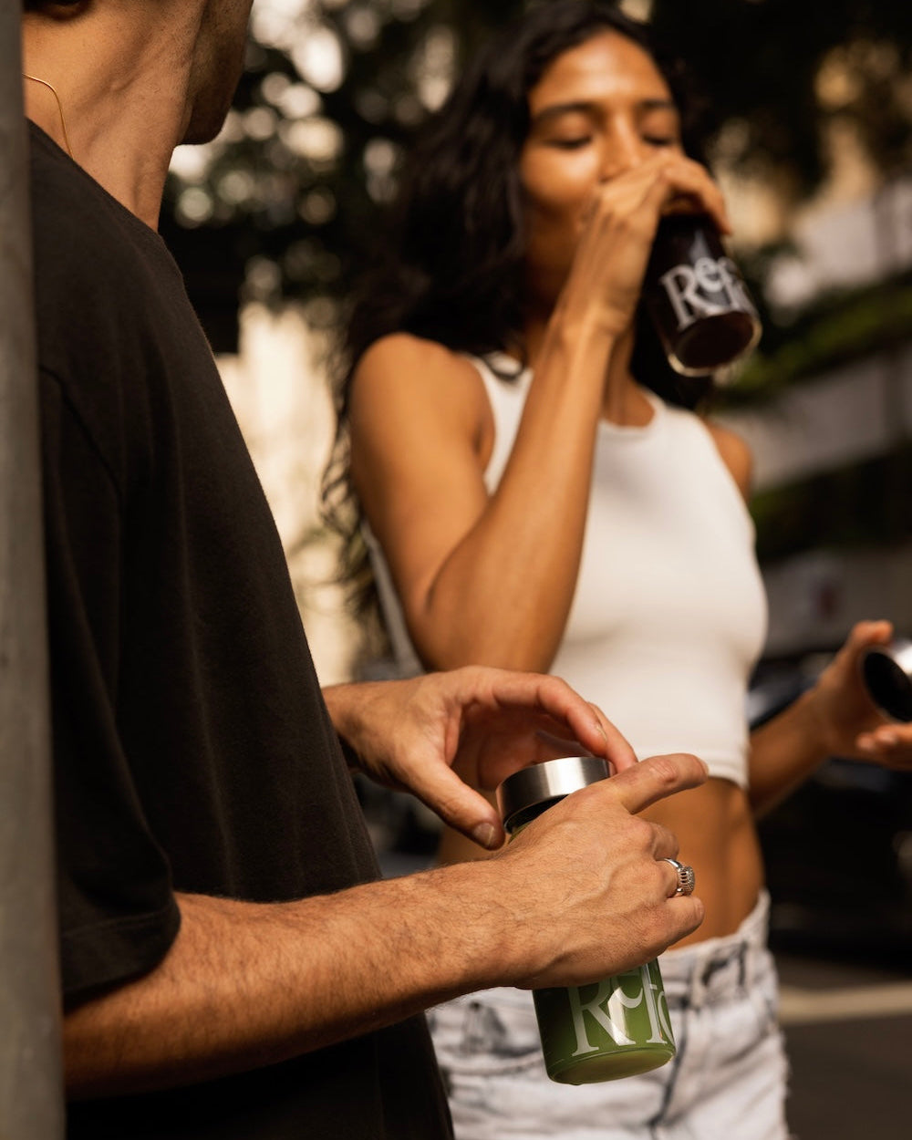 Person holding a green bottle with another person drinking from a red bottle in the background.